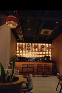 The image is of a cafe and shows a bar with lamps on top hanging from the ceiling and 4 chairs.